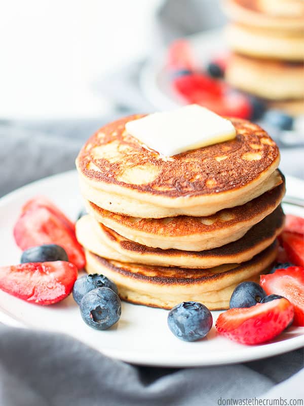 Overview of a stack of pancakes, with butter and syrup on top, surrounded with blueberries and strawberries. A fork sits on the round white plate with a bite ready to eat. Text overlay Paleo Almond Flour Pancakes.