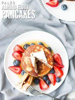 Overview of a stack of pancakes, with butter and syrup on top, surrounded with blueberries and strawberries. A fork sits on the round white plate with a bite ready to eat. Text overlay Paleo Almond Flour Pancakes.