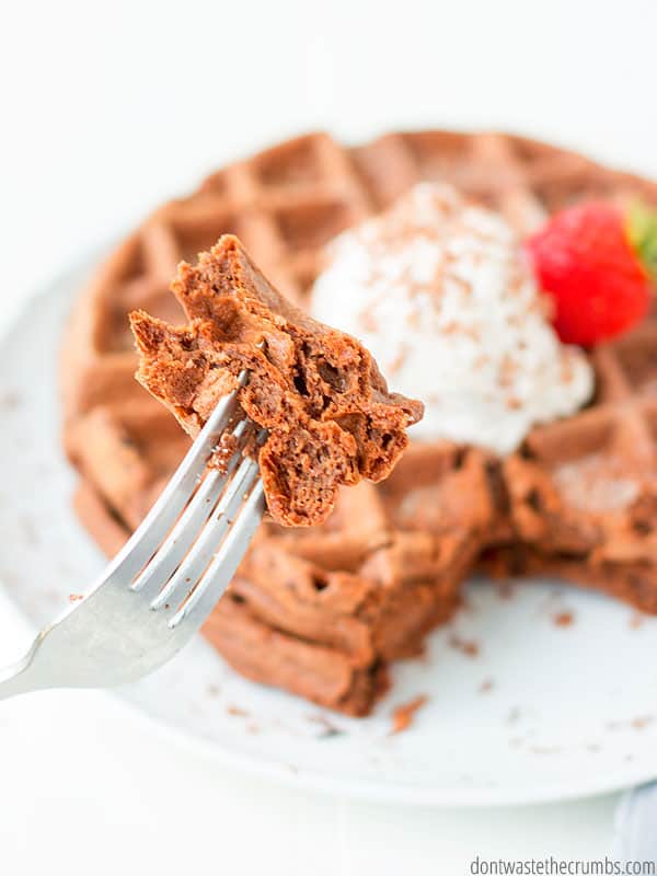 Close up of a bite of chocolate waffle on a fork, with the waffles on a plate in the background.