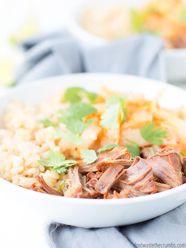 White bowl filled with rice, shredded beef, kimchi and garnished with cilantro, with a sliced lime wedge on a gray kitchen towel. Text overlay Korean Beef Bowls. 