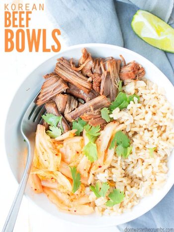 White bowl filled with rice, shredded beef, kimchi and garnished with cilantro, with a sliced lime wedge on a gray kitchen towel. Text overlay Korean Beef Bowls.