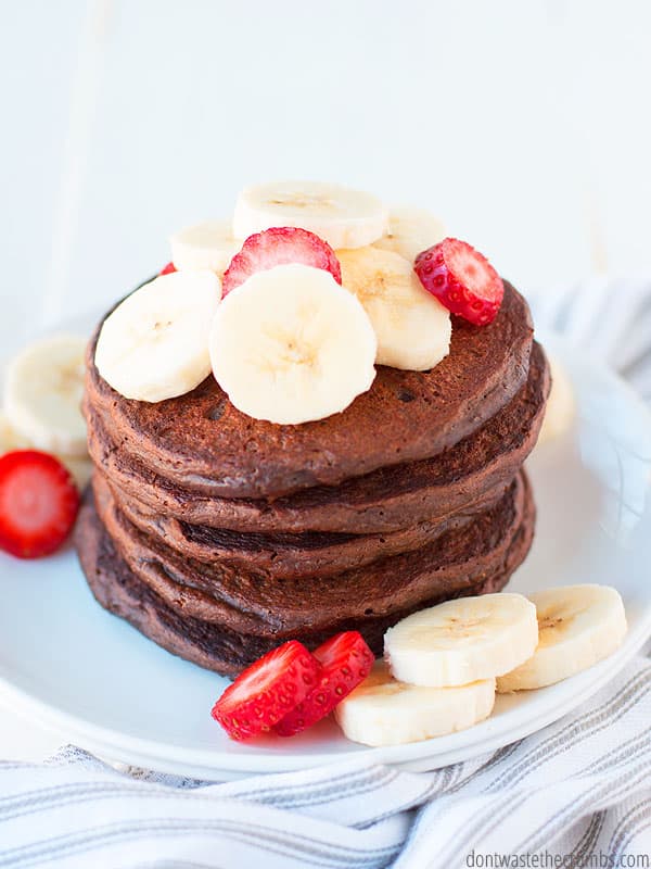 Tall stack of chocolate pancakes on a plate, topped with sliced bananas and strawberries.