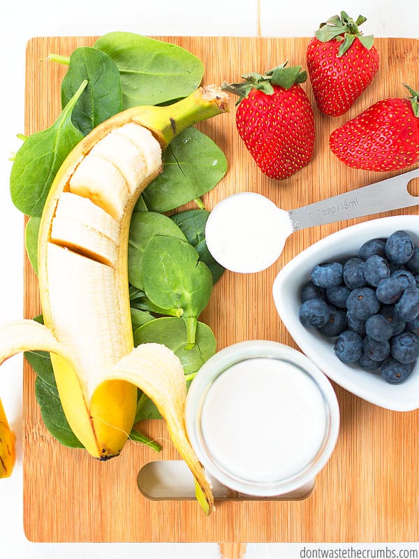 Banana (half in slices), spinach leaves, container of yogurt, bowl of blueberries, measuring cup of yogurt, fresh strawberries on a cutting board.