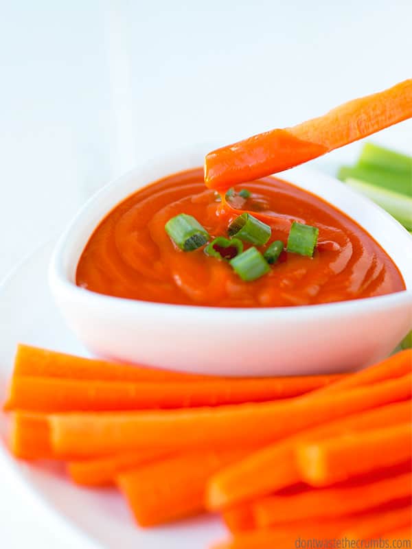 Sliced carrot sticks are being dipped into a bowl of homemade buffalo sauce(with chopped chives on top) and there are also celery sticks on the plate platter.