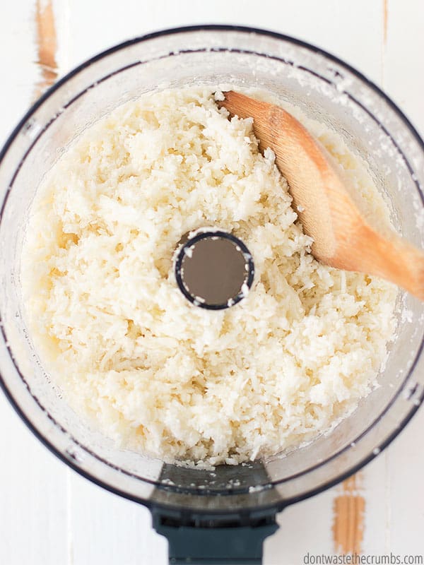 An overview shot of cauliflower rice in food processor with a wooden spoon.