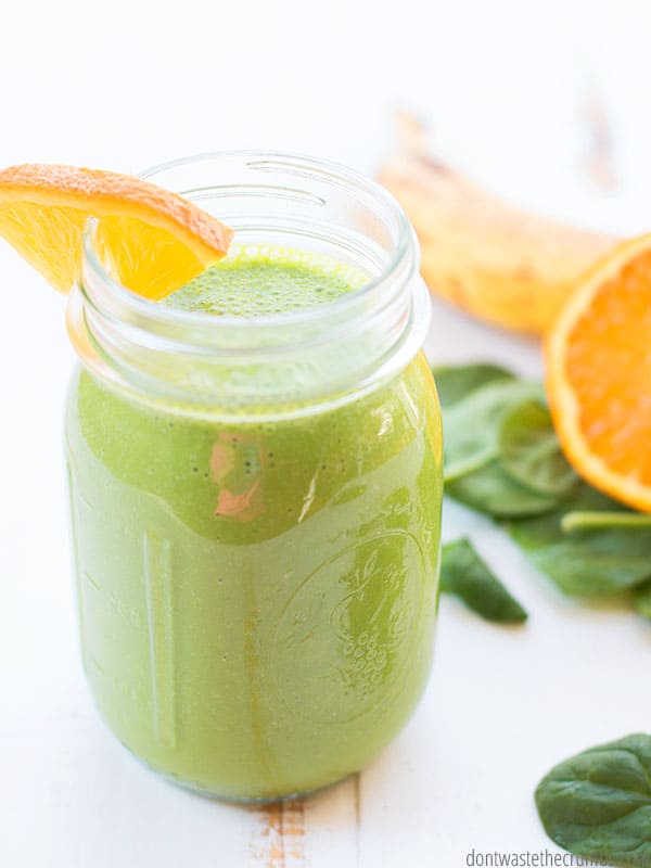 Green Smoothie in a mason jar, with an orange slice on top. It's surrounded by spinach leaves and another orange slice. 