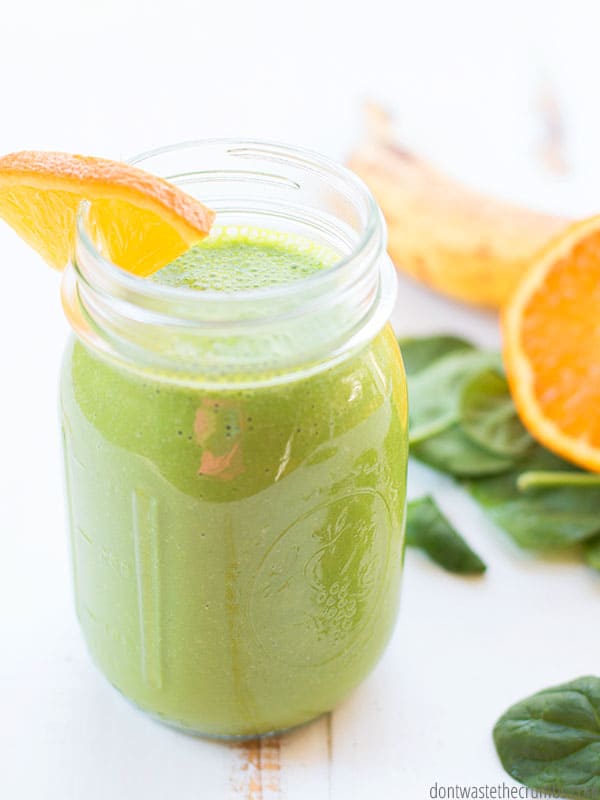 Large glass mason jar filled with green smoothie and a wedge of orange on the rim of the glass. Sliced orange and spinach leaves in the background.