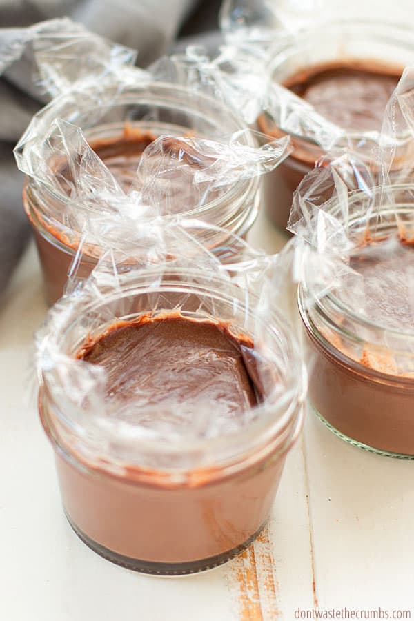 Homemade chocolate pudding inside four small mason jars. Plastic wrap is on top of each jar, covering the pudding. 