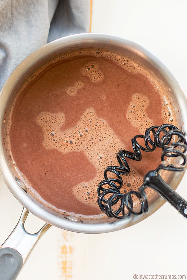 A black flat whisk stirring melted chocolate chips in milk inside a saucepan. A grey towel is in the background. 