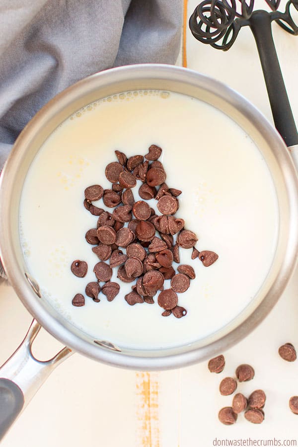 Milk inside a sauce pan, topped with chocolate chips. A Flat whisk and a grey towel are in the background. 