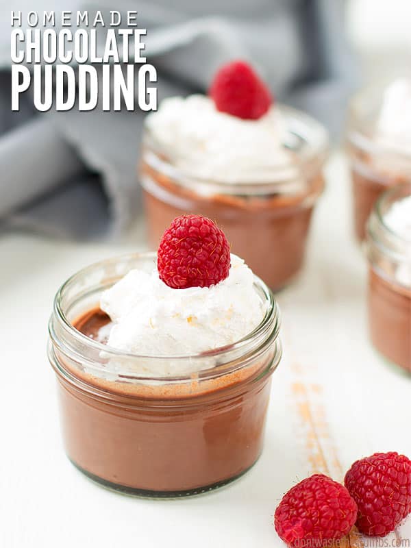 Homemade Chocolate Pudding in a small mason jar, topped with whipped cream and a raspberry. In the foreground are two raspberries on the table. In the background are more jars of pudding. The text overlay says, "Homemade Chocolate Pudding."