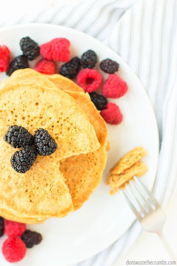 A fork holds a bite of sourdough pancakes, on the side of a white plate. There is a sliced stack of pancakes with fresh berries drizzled all over the plate.