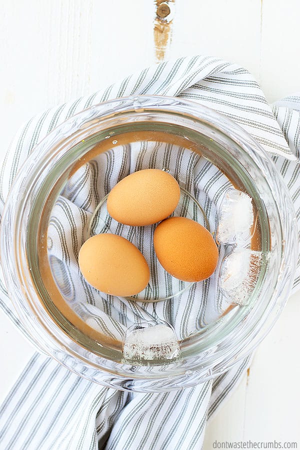 Three boiled eggs sit in a glass bowl, encased in water and ice cubes.