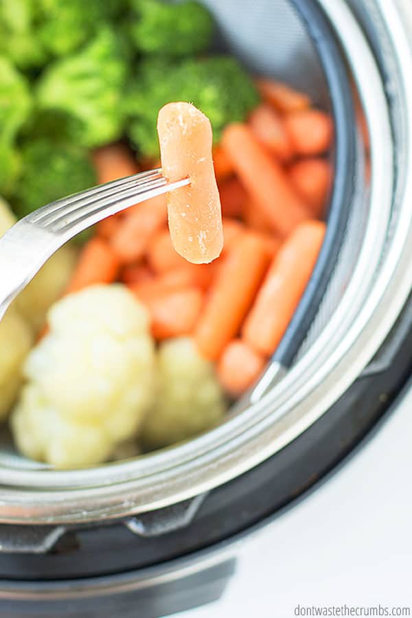 A baby carrot on a fork, with an instant pot in the background with a steamer basket containing cauliflower, broccoli, and baby carrots