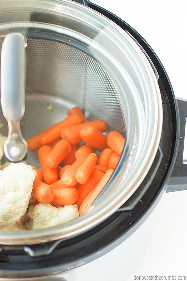 An instant pot with cauliflower and carrots in a steamer basket, covered by a glass lid