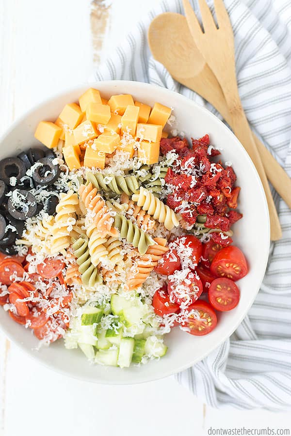 Overhead view of a big bowl of freshly mixed pasta salad, finished with parmesan cheese. Wood spoon and fork nearby.