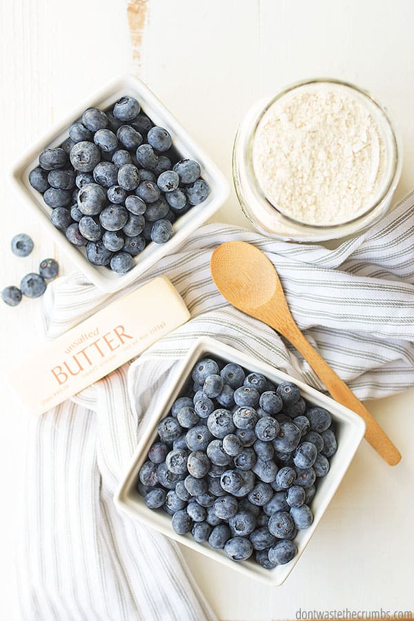 Blueberries in two white bowls and a stick of butter and a cup of flour on a table next to a wood spoon.