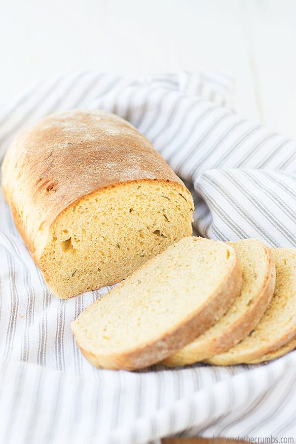 Rosemary olive oil bread loaf is cut in half and one half is cut into slices on a table cloth.