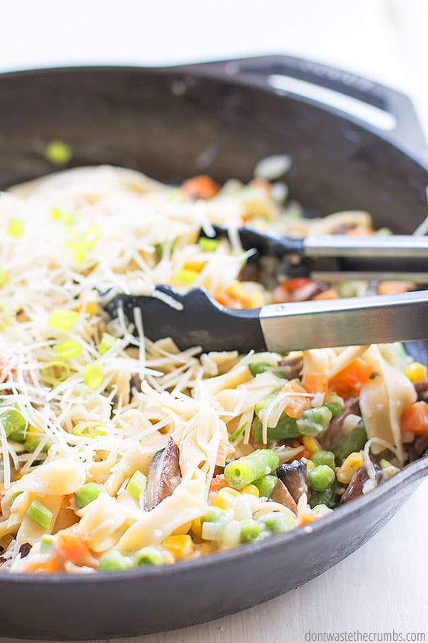 Pasta primavera in a cast iron skillet and tong mixing the ingredients and shredded cheese together.