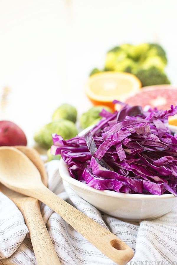 Shredded cabbage in a white bowl and other vegetables in the background on a table. Two wooden spoons next to the white bowl.