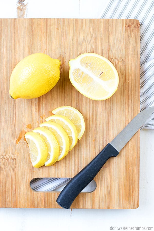 A wooden cutting board is pictured with two lemons. One lemon is whole, and the other is sliced in half. One of the halves is sliced into wedges. There is a paring knife on the cutting board with a black handle.  