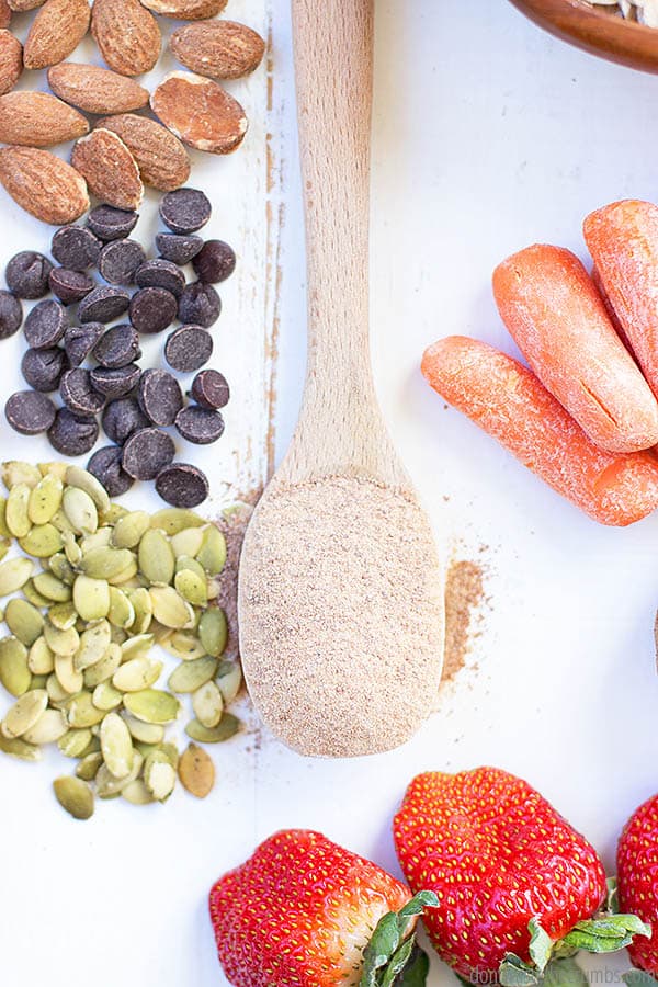 save on food: A pile of nuts, a pile of chocolate chips, and a pile of seeds on the left side of a table. There is a wooden spoon in the middle of the table. On the right side there is a pile of baby carrots.