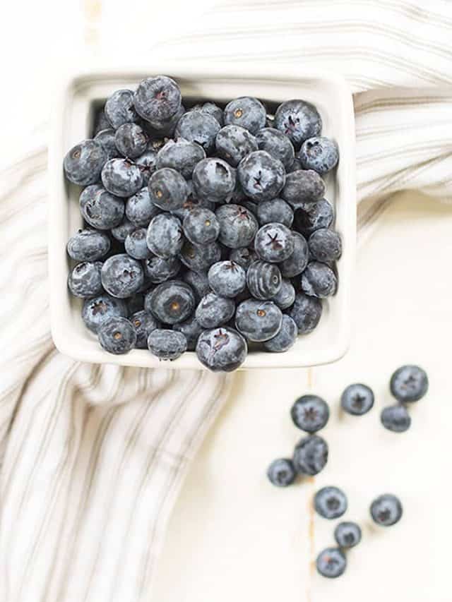 White square dish full of blueberries on top of a cloth and a few blueberries laying next to it