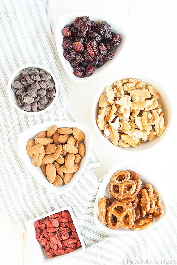 Bowl of chocolate chips, bowl of dried cranberries, bowl of walnuts, bowl of almonds, bowl of pretzels, bowl of pepitas.