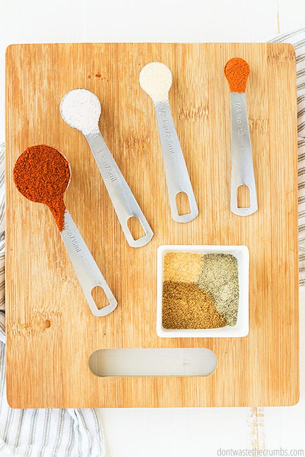 Spices in different measuring spoons and a square bowl on top of a wooden cutting board