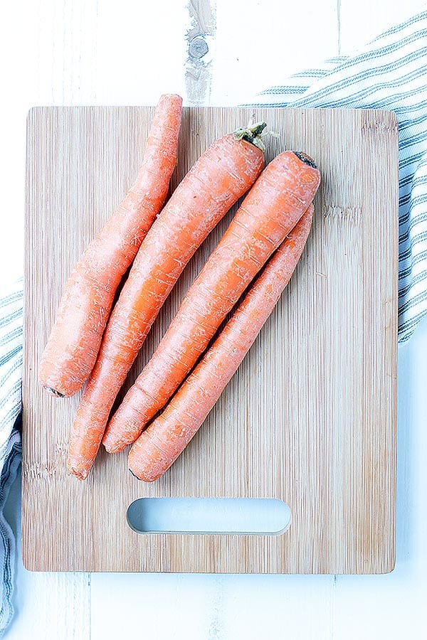 Four unpeeled, untrimmed, large carrots sit on top of a wooden cutting board.