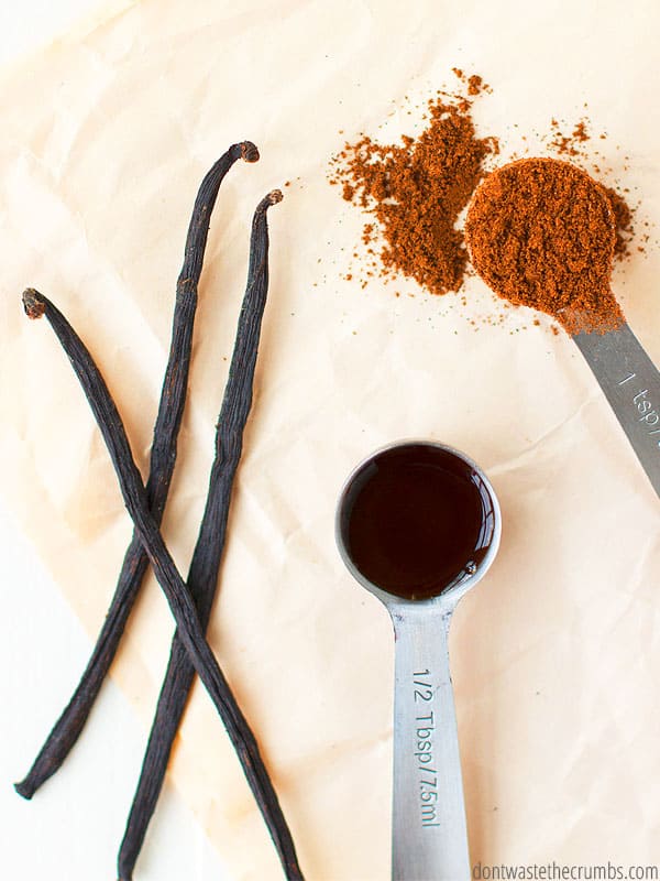 Three vanilla beans on a cream cloth to the left next to a measuring spoon of vanilla extract in the middle and a measuring spoon of vanilla bean powder on the right.