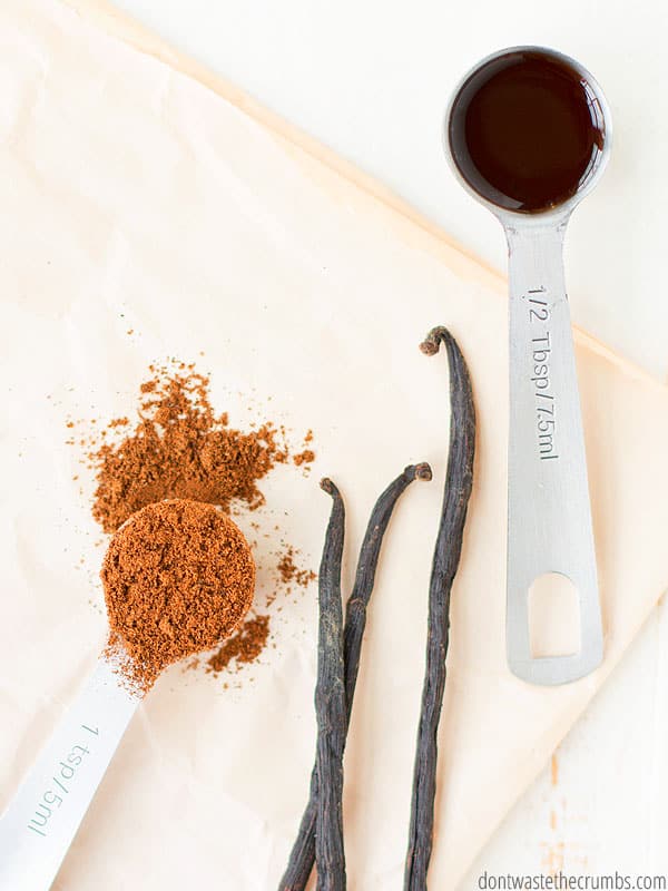 A casually laid out assortment of vanilla  products including vanilla bean powder on the right, The tops of three vanilla beans in the middle, and a measuring spoon of vanilla extract on the right.