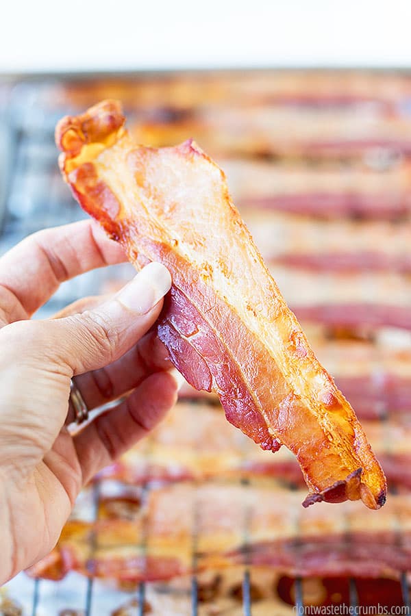 A hand holding a cooked slice of bacon in front of the blurred out pan that contains the rest of the bacon.