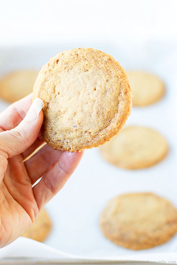 A well manicured hand holding a whole cookie. Other cookies are in the background on parchment paper and a cookie sheet.