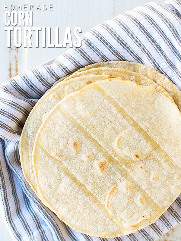 A stack of golden brown corn tortillas on top of a gray and white striped towel. The text over lay says, "Homemade Corn Tortillas".