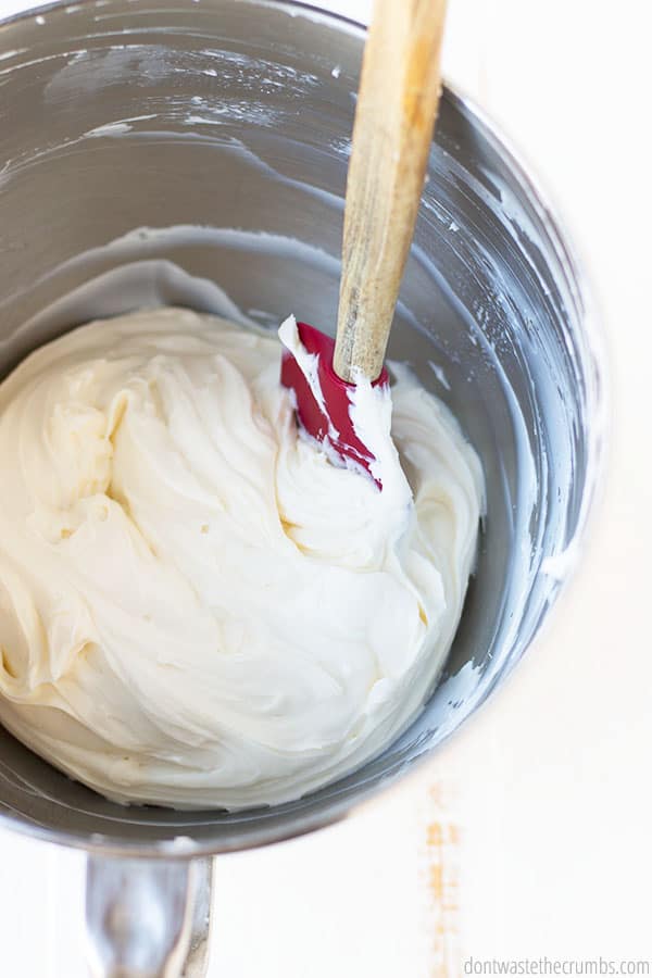 A metal mixing bowl full of white, creamy, and fluffy delicious filling which is being smoothed with a red spatula. 