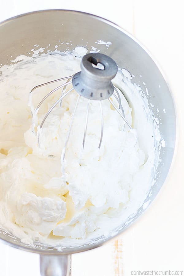 Whipped cheesecake filling in a large metal mixing bowl. A stand mixer whisk is in the bowl making my want to scoop it up and lick off the delightful filling.