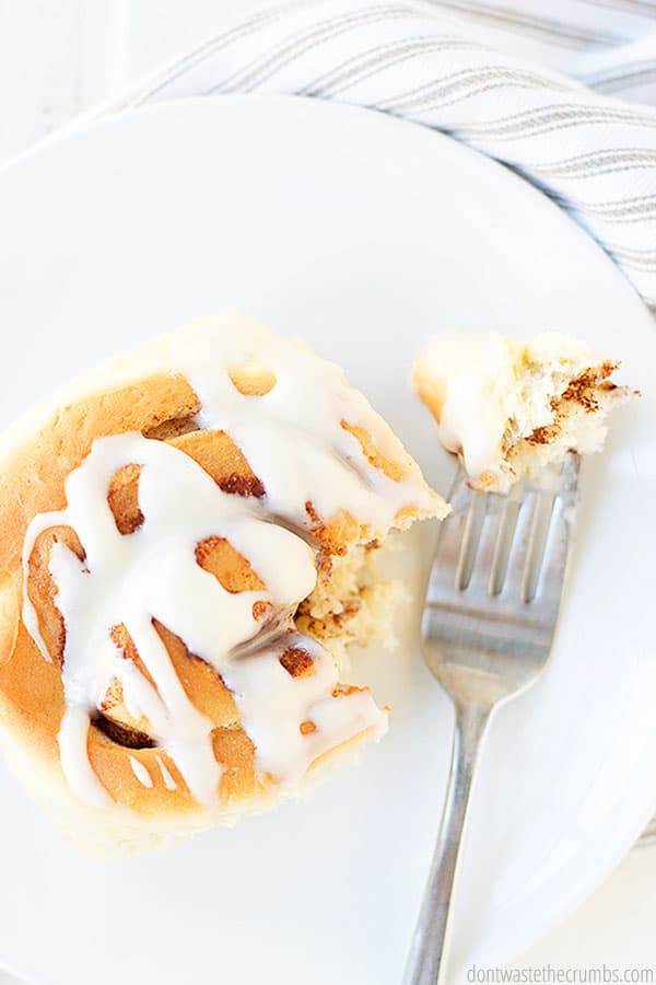 A fork with a bite-sized piece of cinnamon roll, next to a warm cinnamon bun, smothered in fresh homemade vanilla glaze. 