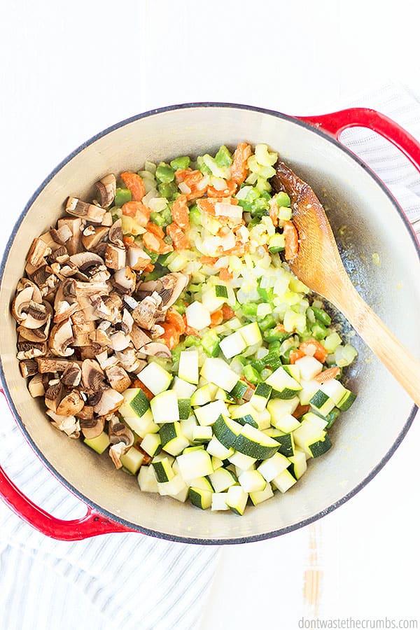 A stock pot filled with diced vegetables such as carrots, green bell peppers, onions, mushrooms and zucchini. A wooden spoon is being used to mix the vegetables with roux.