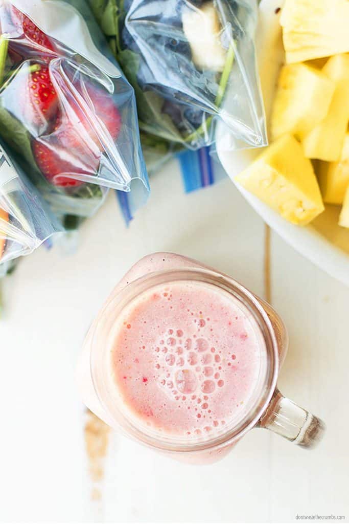 A freshly blended smoothie in a glass mason jar sits beside three smoothie packs and a fresh bowl of pineapple.