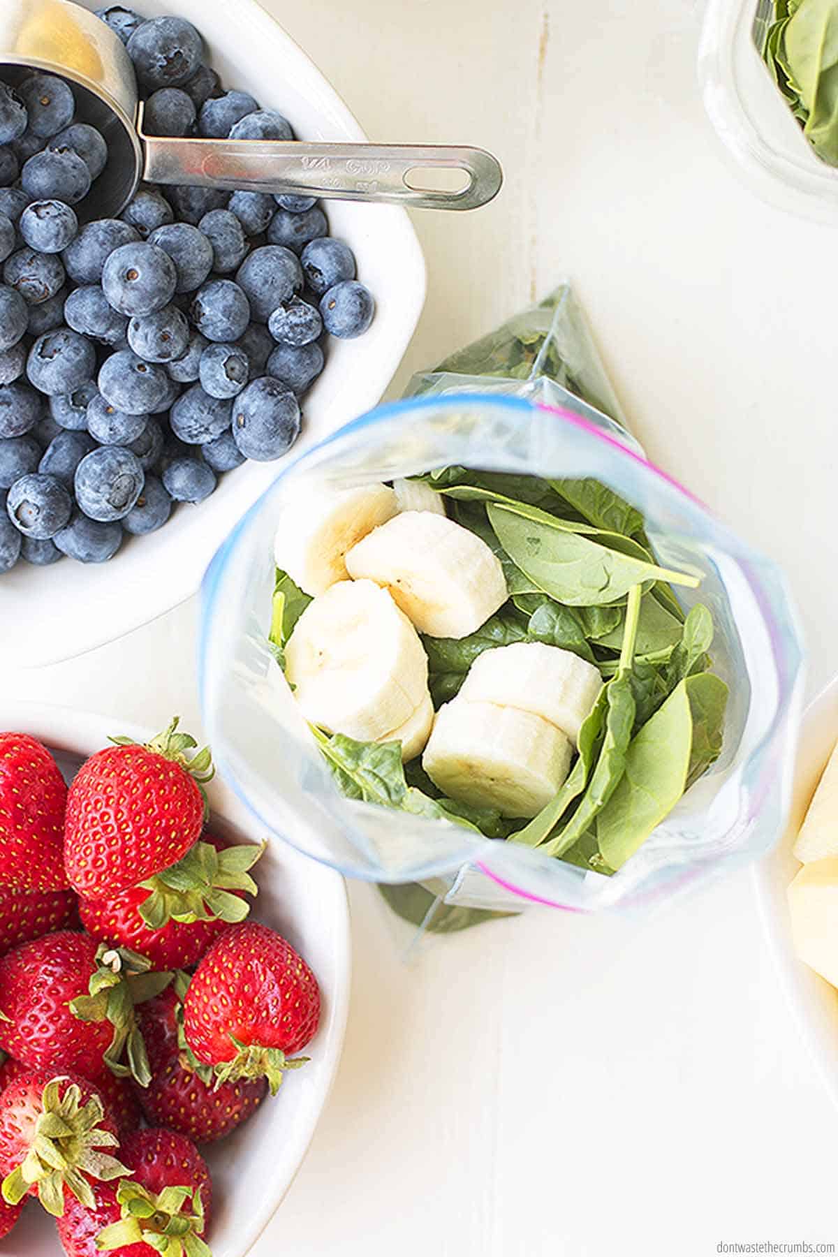 A freezer bag is being filled with fresh leafy greens and bananas. A bowl of blueberries has a stainless steel scoop inside of it. and there is a fresh bowl of strawberries as well.  