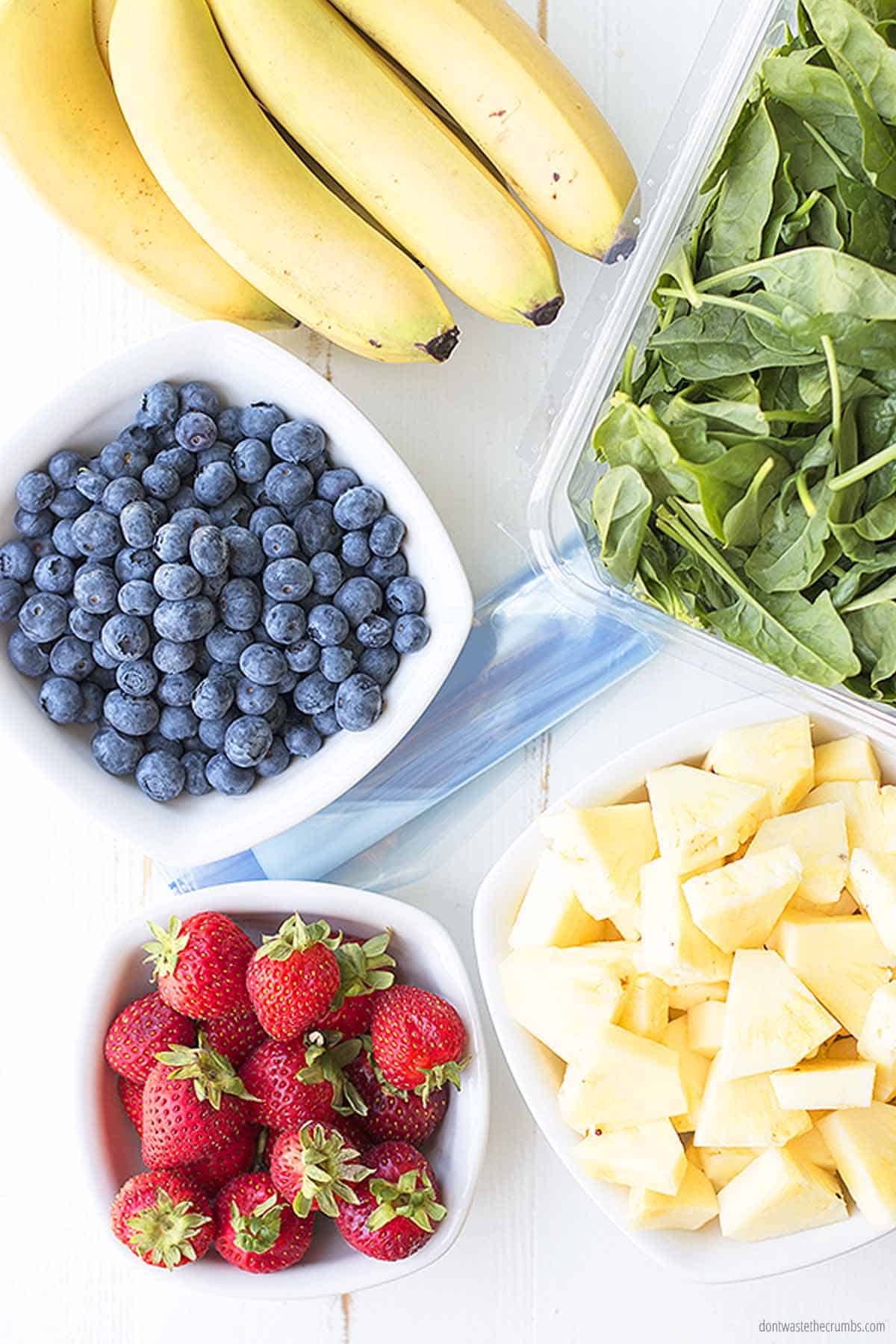 Five separate fresh fruit ingredients ready to be added to smoothie packs. There is a bowl of blueberries, strawberries, pineapple, and kale. There is also an unpeeled bunch of bananas.