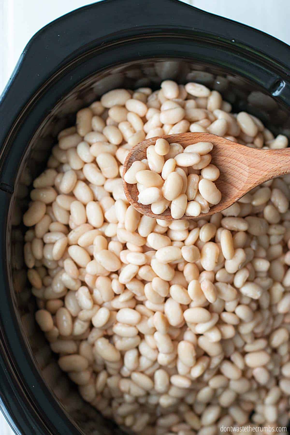 A black slow cooker is filled to the top with cooked white beans. A wooden spoon is serving a heaping pile of beans.