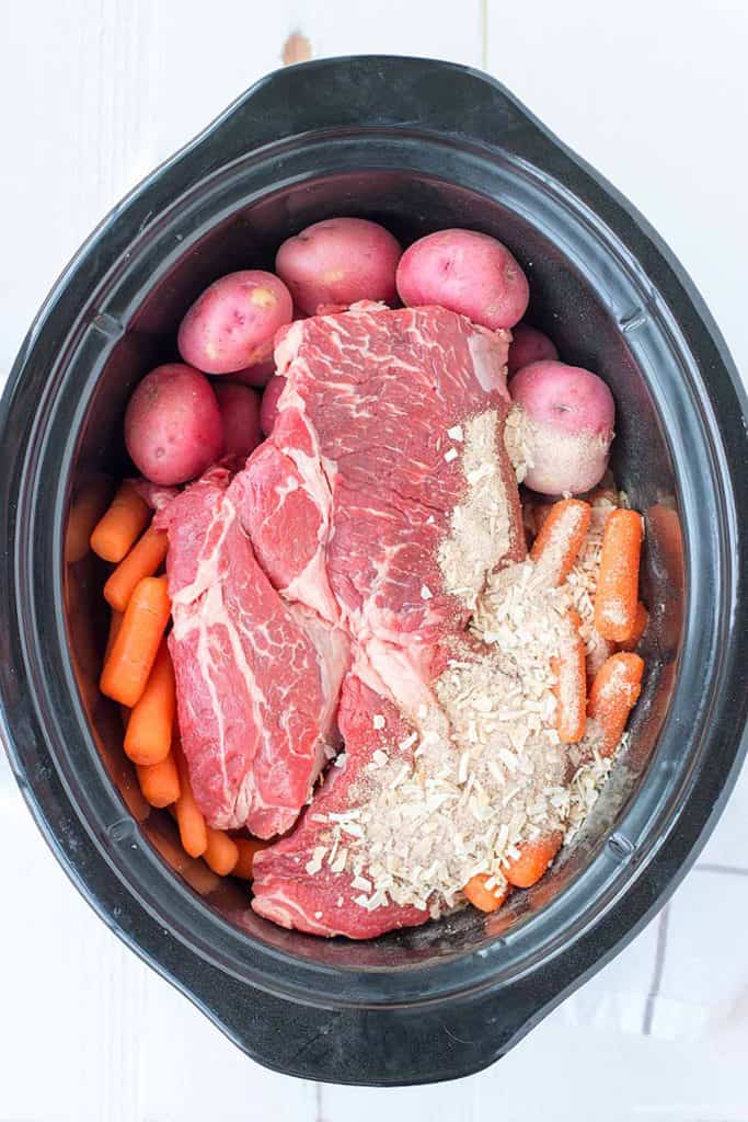 A black oval crock pot is filled with uncooked chuck roast, and fresh carrots  & potatoes. Topped with homemade dry onion soup mix. 