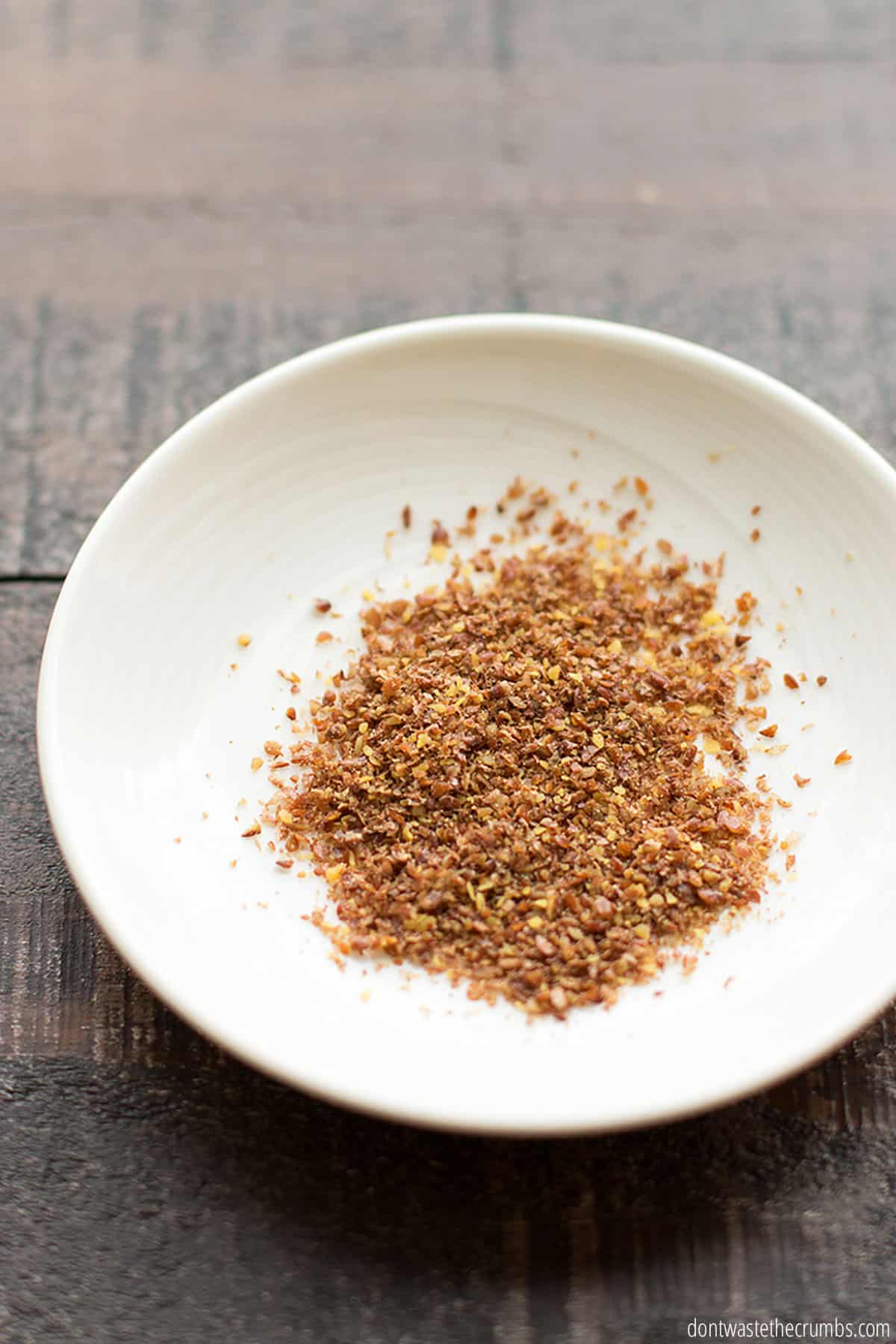 A small white plate on a wooden table with 1 tablespoon of ground flax seeds. 