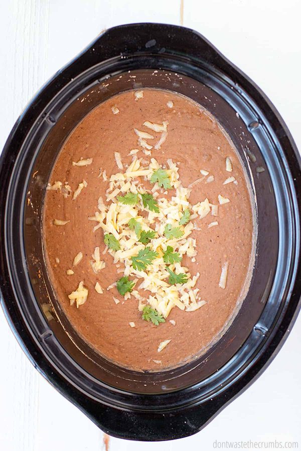 This crock pot refried beans dish is better than the store bought canned version. Topped with shredded cheese and cilantro. Try this easy recipe today!