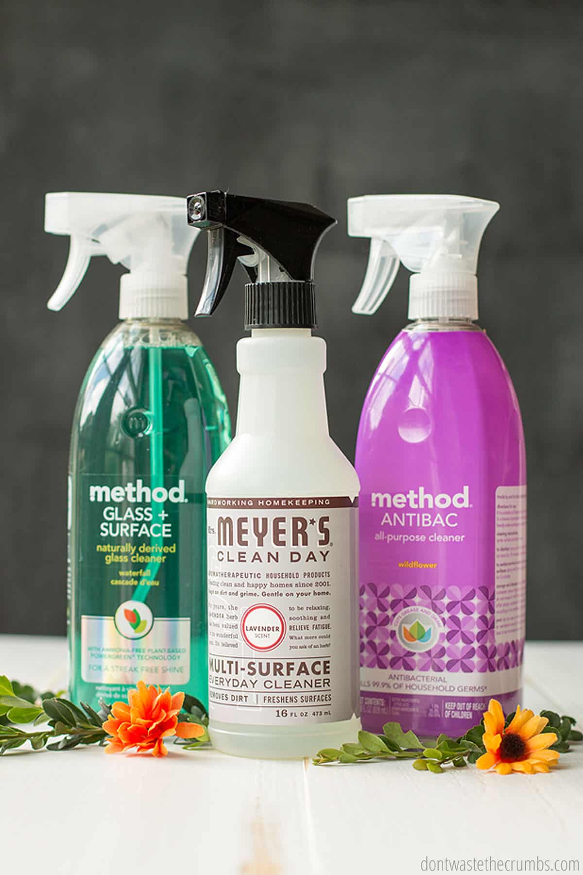 Three bottles of spray cleaning products on a table, set in a row beside some orange and yellow flowers. There is a glass cleaner, an all-purpose cleaner, and an antibacterial surface cleaner. 