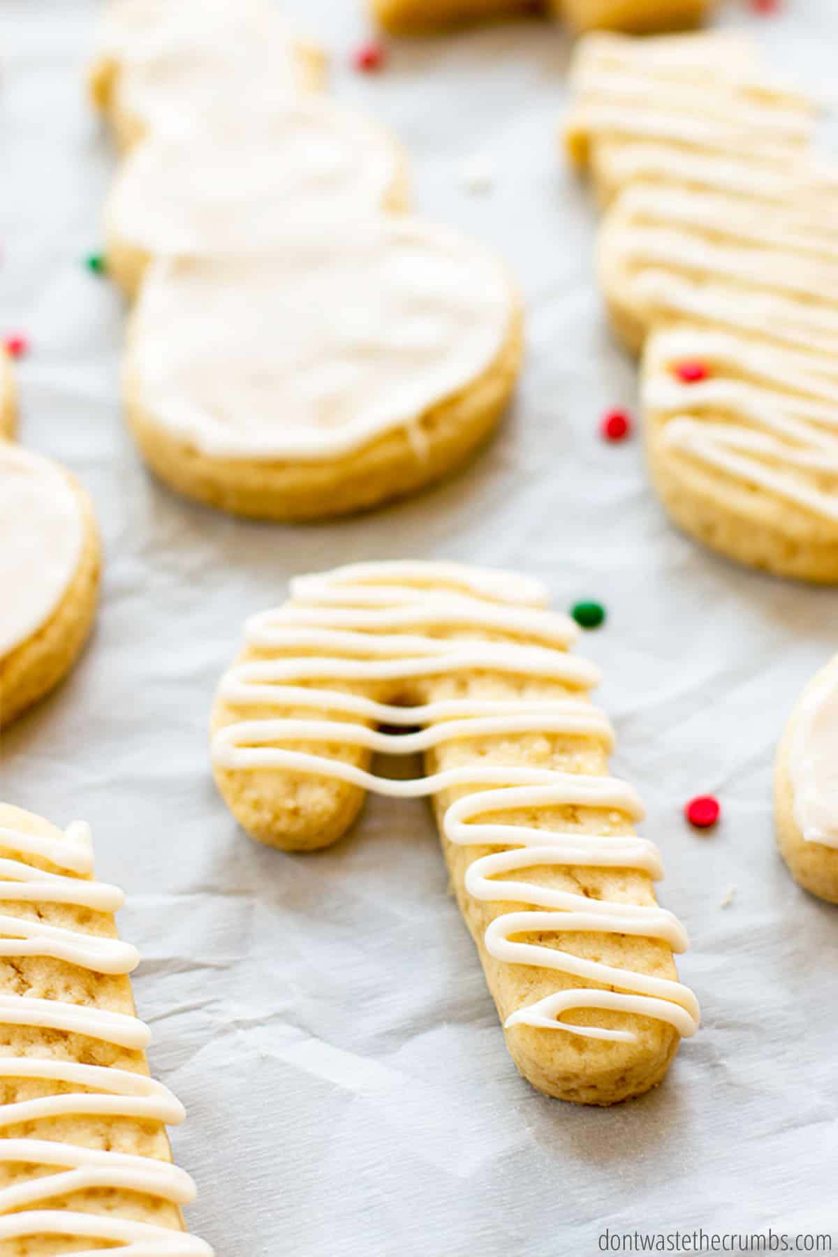 A cookie in the shape of a cane is featured with a zigzag of white icing. In the background are other freshly iced cookies in the shapes of snowmen.