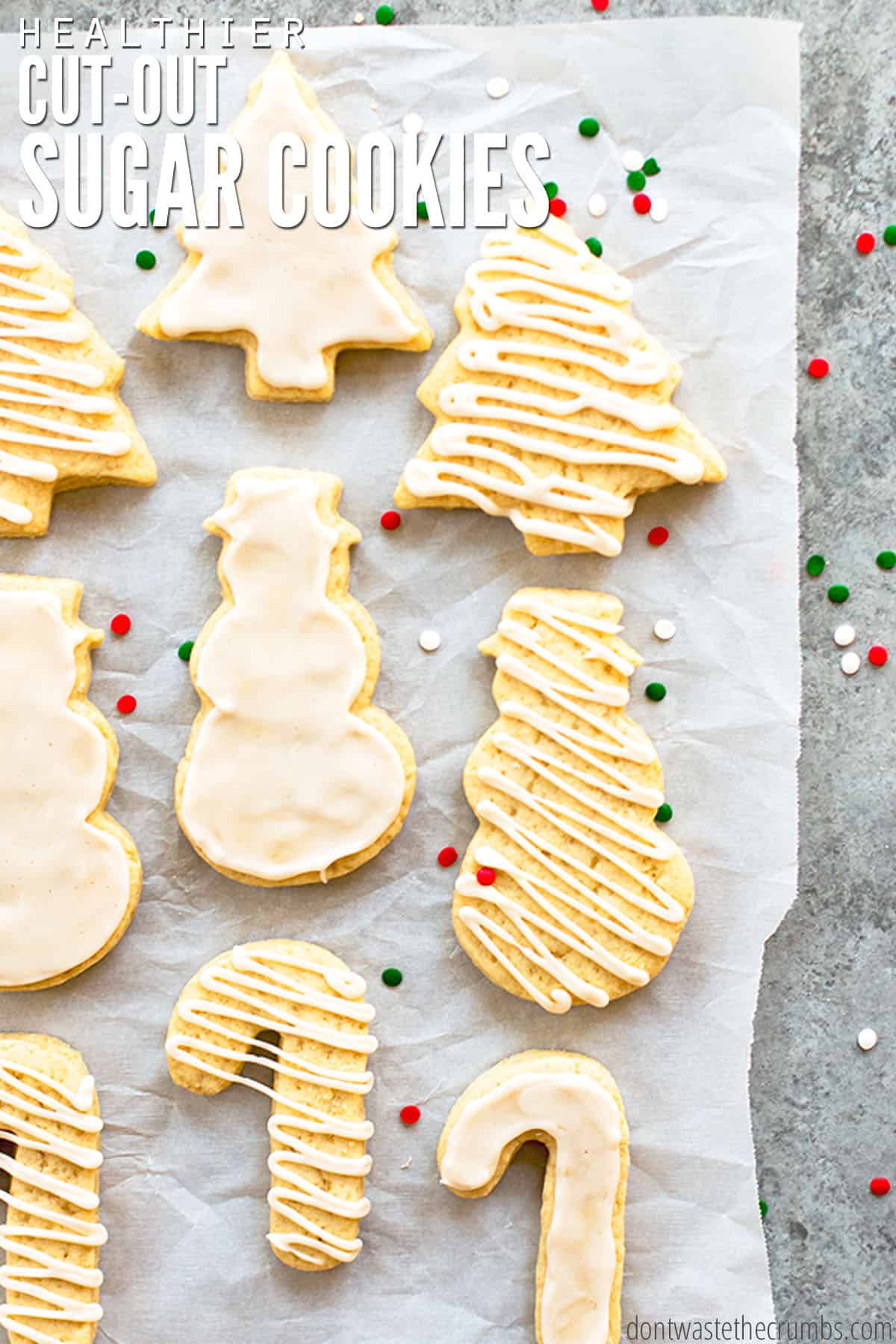 Nine freshly baked sugar Christmas cookies with white icing are spread out on a piece of parchment paper. There are red, green, and white round sprinkles all around the cookies. The text overlay reads, "Healthier Sugar Cookie Recipe."