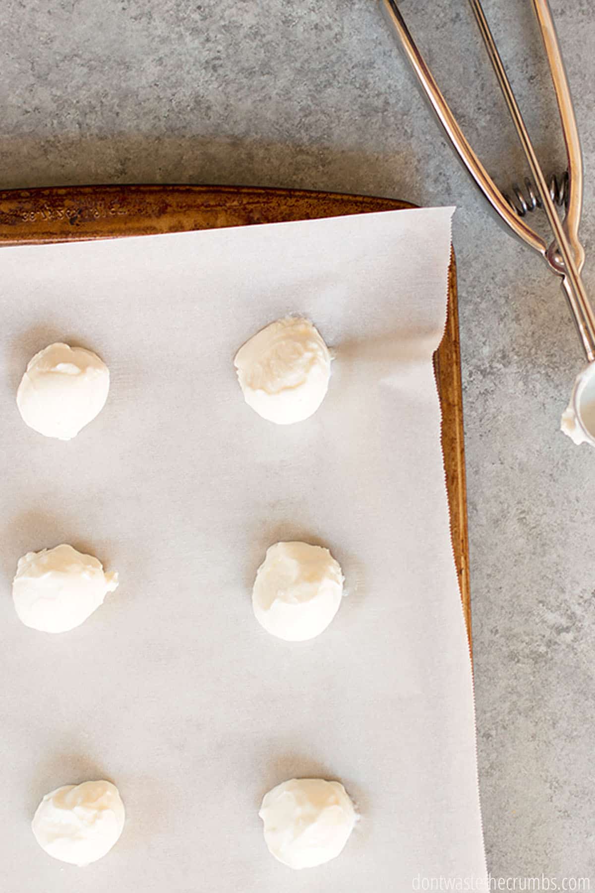 Six freshly scooped balls of yogurt ready to be frozen on a sheet pan with a piece of parchment paper. There is a yogurt scoop beside the sheet pan.